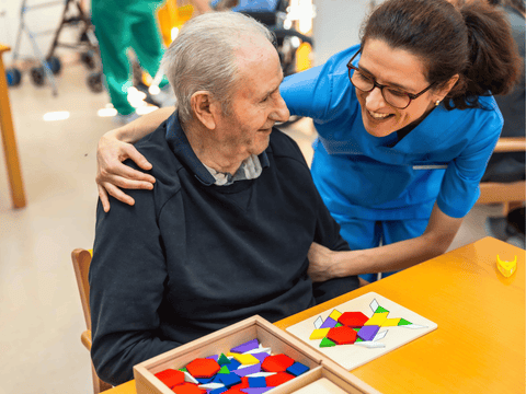 A female caregiver in blue scrubs smiles warmly, with her arm around an elderly man who is sitting at a table with colorful puzzle pieces, illustrating the compassionate care needed when using oral care products for dementia patients