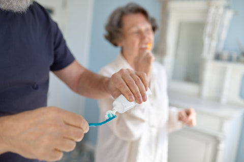 Senior couple standing side by side, smiling while brushing their teeth together in front of a bathroom mirror—promoting healthy habits in aging.