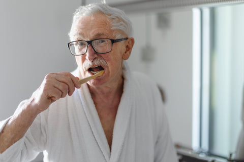 Older man with glasses brushing his teeth in front of a bathroom mirror, focusing on personal oral hygiene.