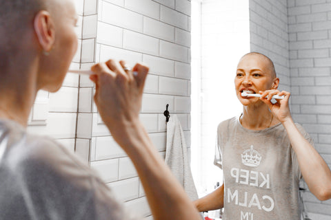 A chemotherapy patient using a soft-bristled electric toothbrush in front of a bathroom mirror, focusing on gentle oral care.