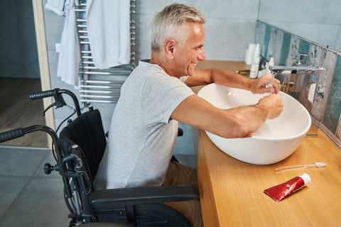 Man in a wheelchair at a bathroom sink preparing to brush his teeth, promoting accessible oral care.