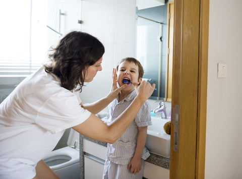 Mother gently brushing her child’s teeth in a bathroom, supporting a calm and caring at-home oral hygiene routine.